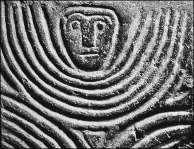 A carved face with concentric circles below, radiating from a central diamond