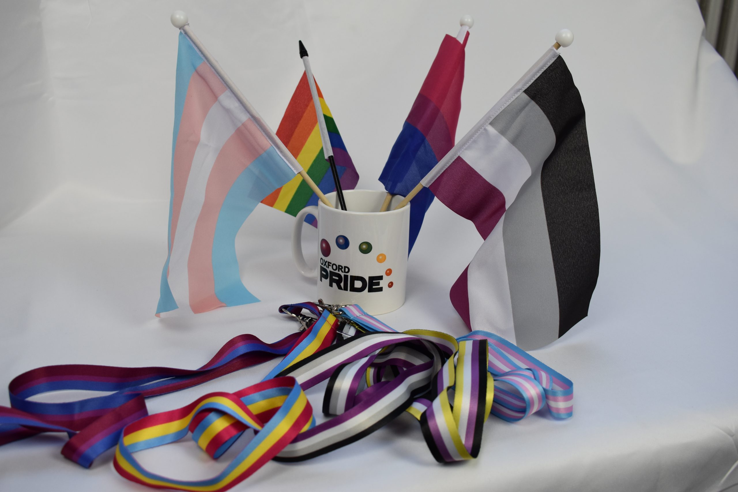A selection of Oxford Pride memorabilia, including a mug with its logo, four mini flags (trans pride, original rainbow, bisexual pride and asexual pride flag) and a selection of lanyards in various LGBTQ+ flag colours