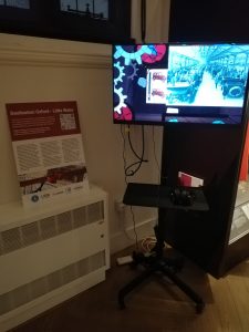 A TV in the museum playing the Destination Oxford - Little Wales film. The screen shows cogs and old pictures of cars. Next to the TV is a poster explaining the project.