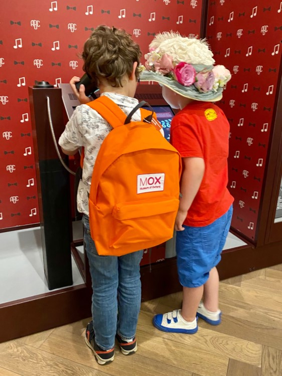 Two children listening at a post. One is wearing an orange backpack and the other is wearing a floral hat