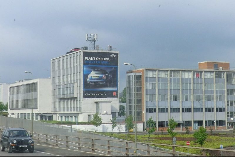 View from a road of a large rectangular white building with an advert for the Oxford Mini Plant