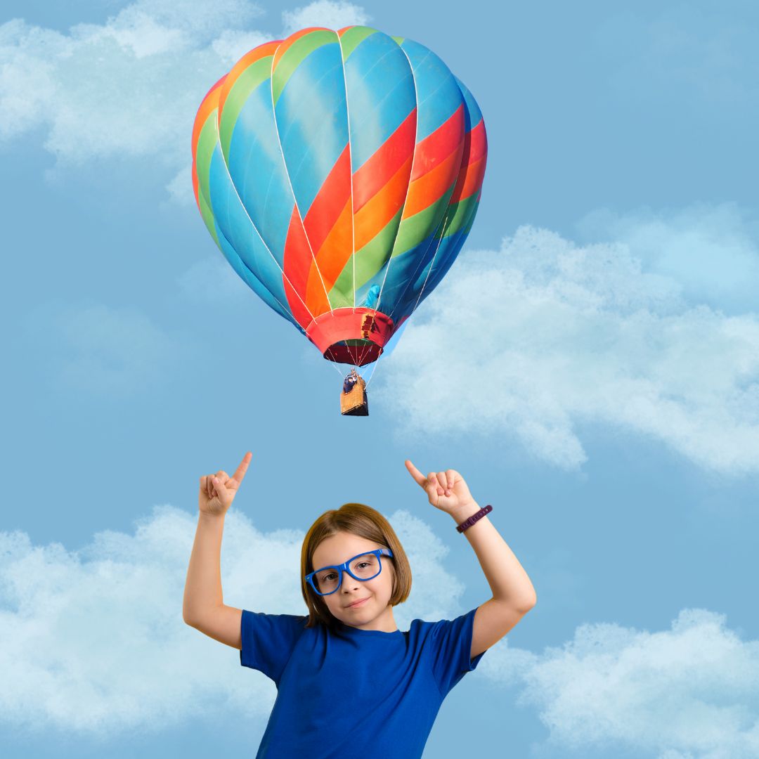 A young child stands in the center and points upwards at a multi-colored hot air balloon, over a blue and white sky background.