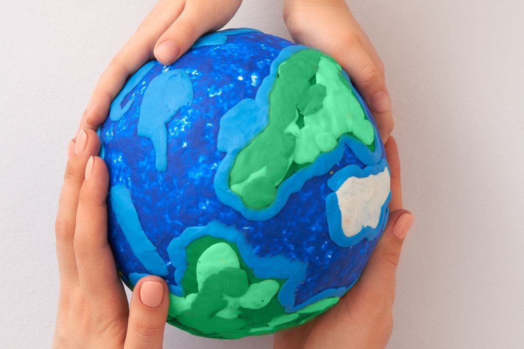 A model of earth made from glue and green clay is held in two pairs of hands, one belonging to an adult and one to a child.