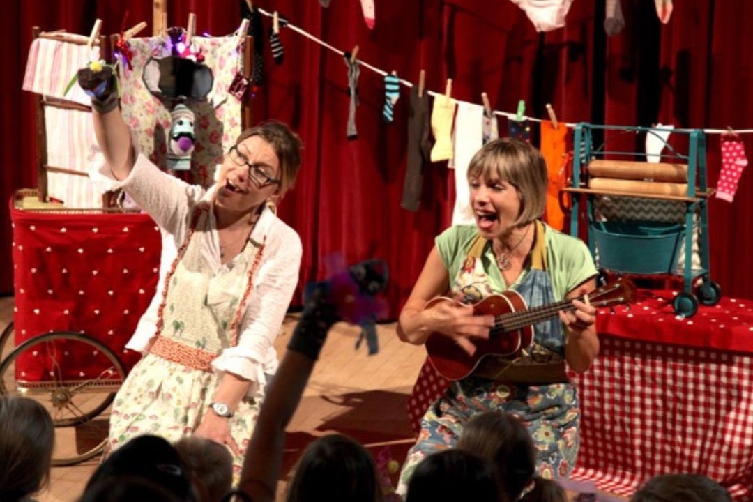 Two women sit in front of an audience of children. One holds a sock puppet, one plays the ukele. In the background is a washing line laden with socks.