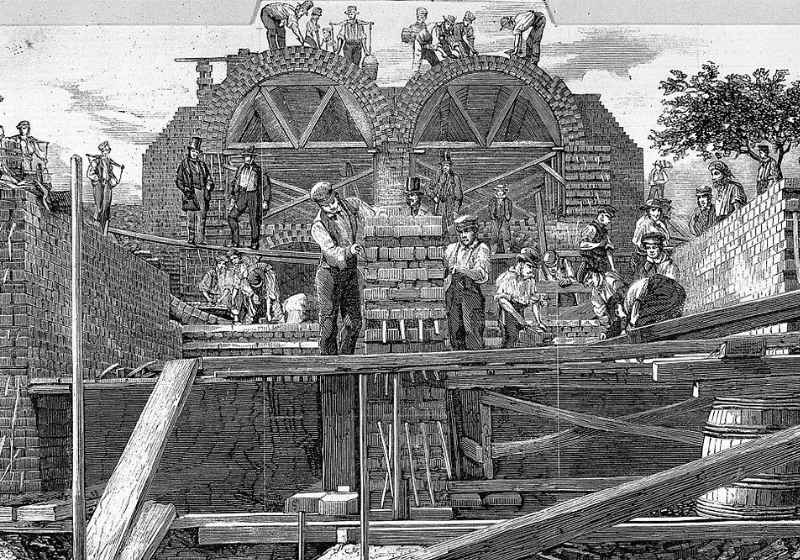 A black-and-white Victorian era drawing showing the construction of sewerage tunnels. Two incomplete brick archways are in the background. In the foreground, two workmen, illustrated with flat caps, white shirts and trousers held up by braces, move a stack of bricks. Other workmen are depicted throughout the image engaged in activities such as brick-laying. Two managers, illustrated wearing top hats and dark blazers, stand on a wooden plank, overseeing the work.