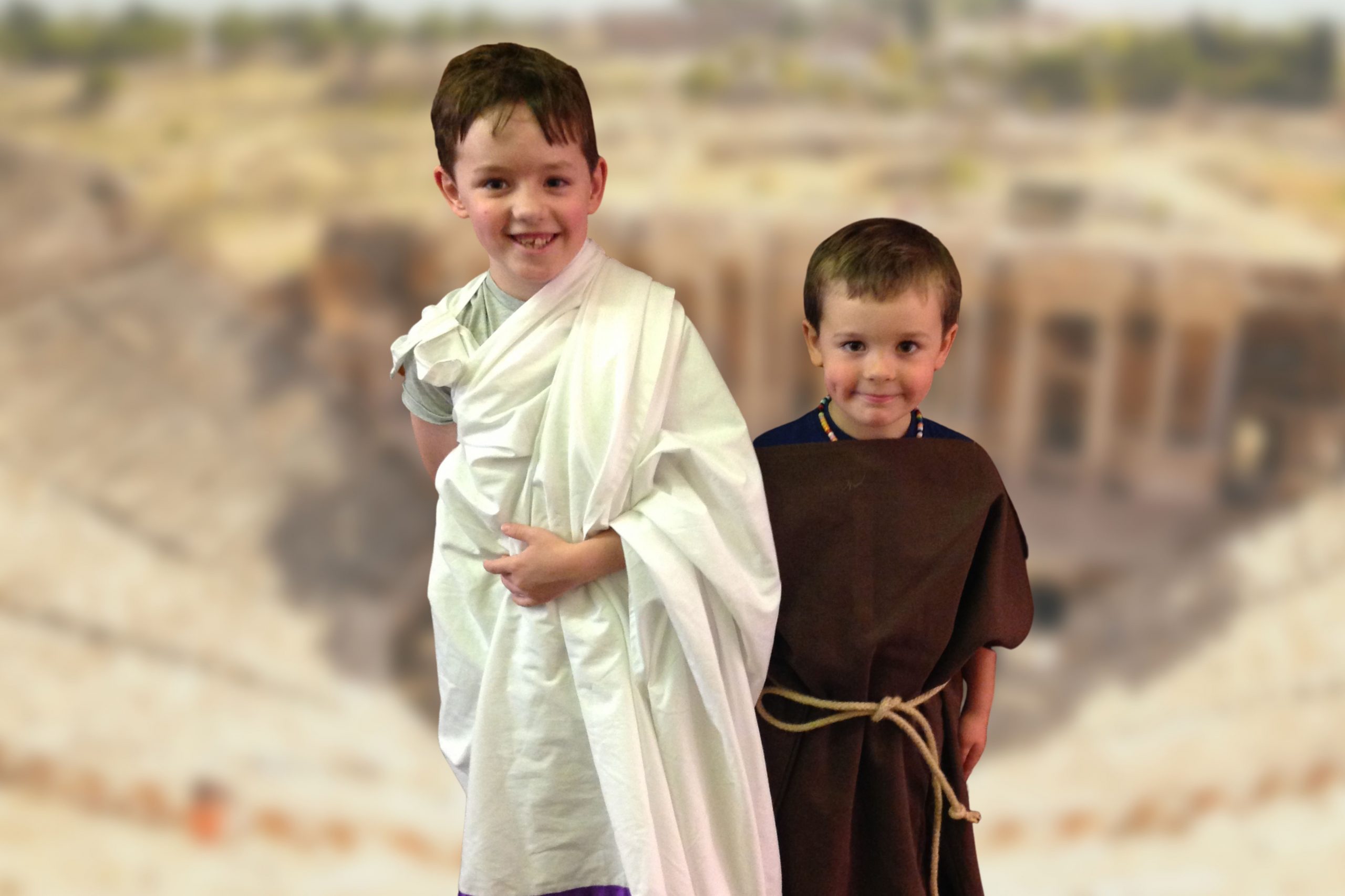 Two small boys, wearing a brown tunic and white toga, stand in front of a blurred image of a colosseum.