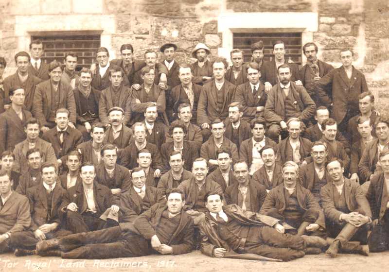 A black and white group photograph of men in working uniforms. The men are sitting and standing in rows in front of a wall with barred windows.