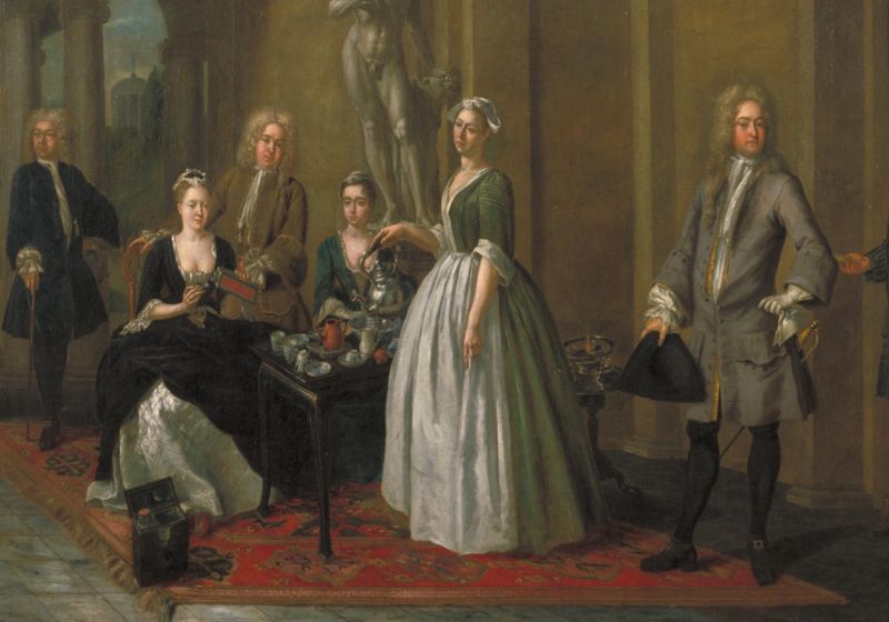 A Georgian era painting depicting a maid in a green smock and white apron serving tea out of a silver tea pot to a wealthy family. The family sit and stand around a table. The women wear dresses with large skirts and the men wear curly wigs.