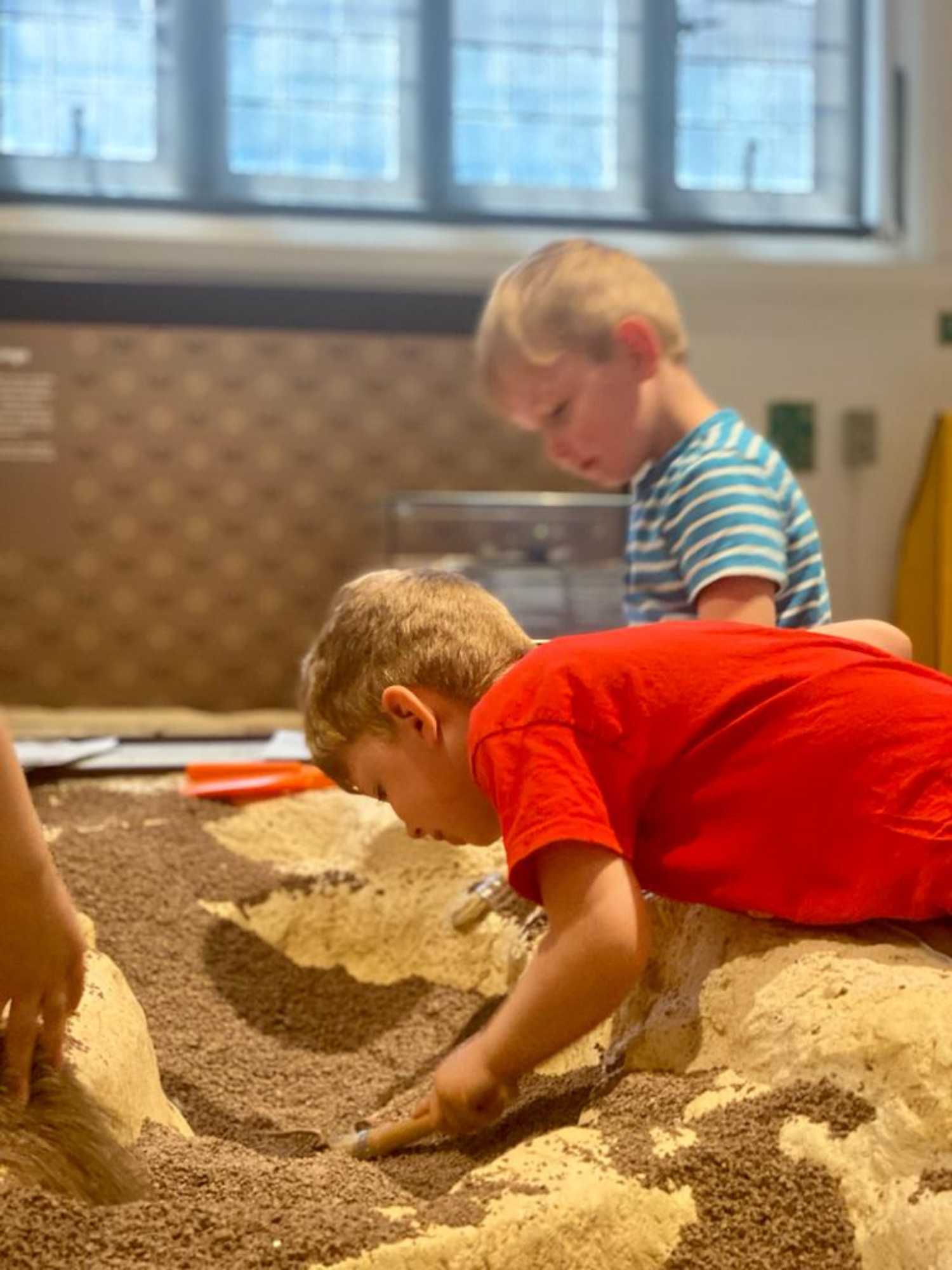 Two children lean over an archaeological play pit and dig through fake mud.