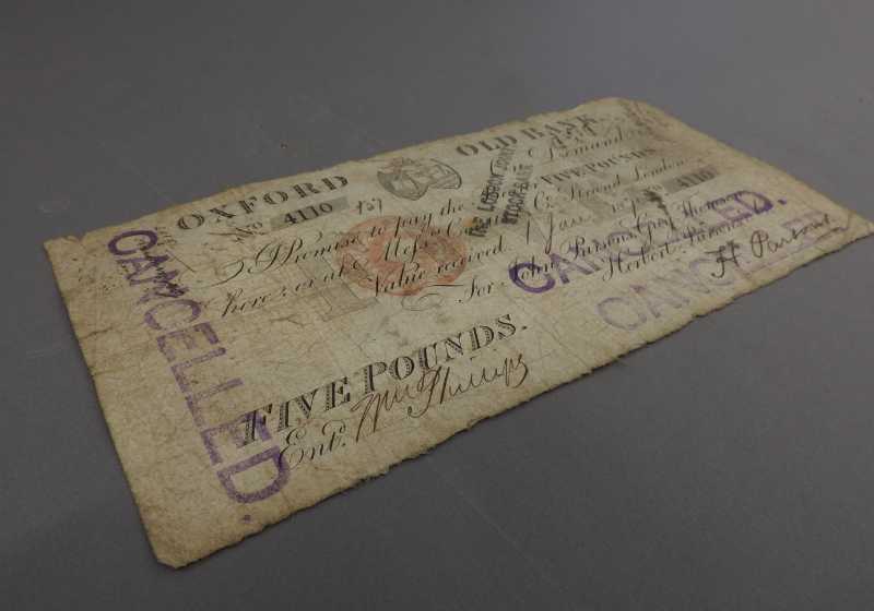 A yellowed old-fashioned five pound bank note from the Oxford Old Bank. Stamped in purple ink across the front is the word 'cancelled'.