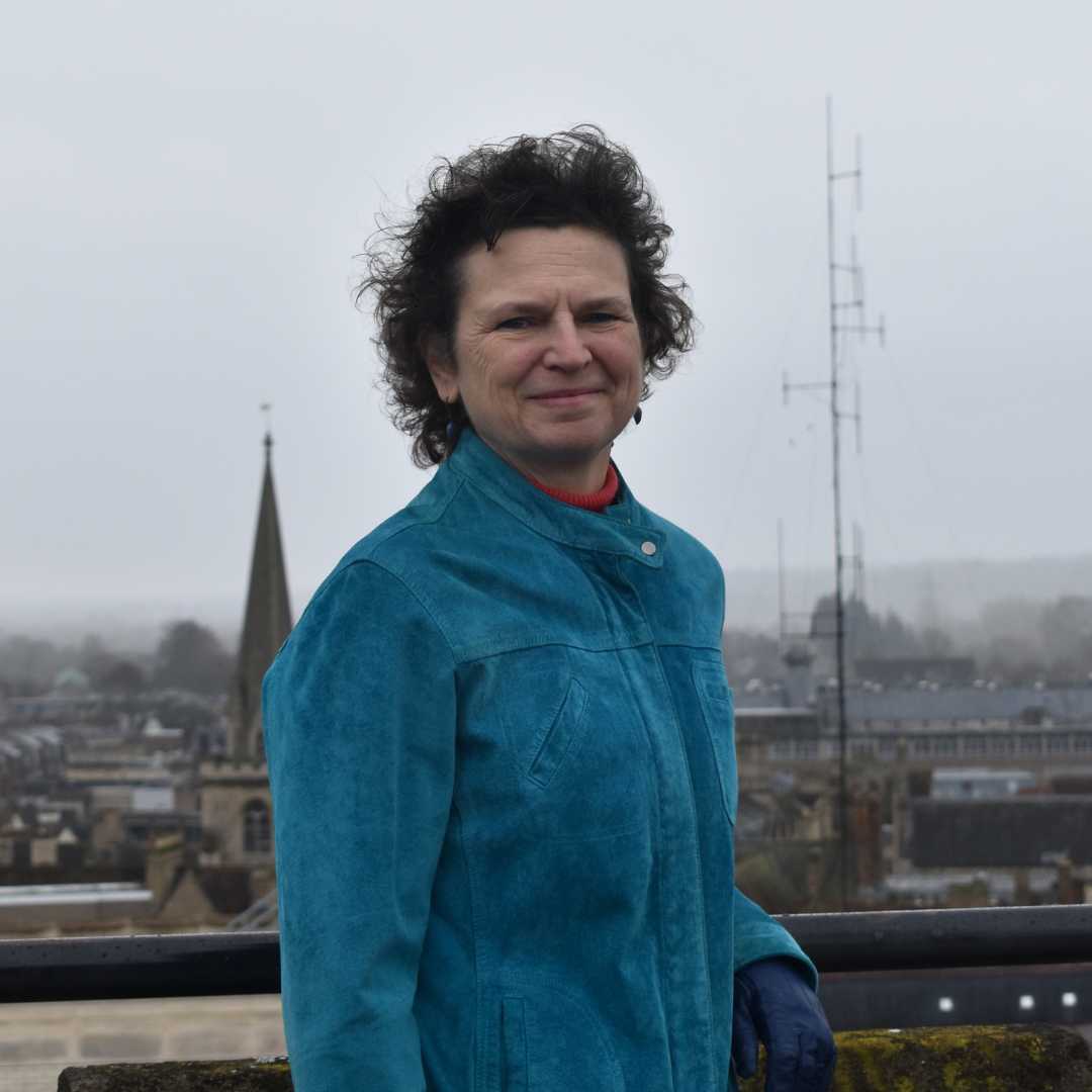 Liz Woolley smiles at the camera as she stands in front of a view of the Oxford skyline on a cloudy day. She has light-toned skin, short curly brown hair and is wearing a teal-coloured jacket.