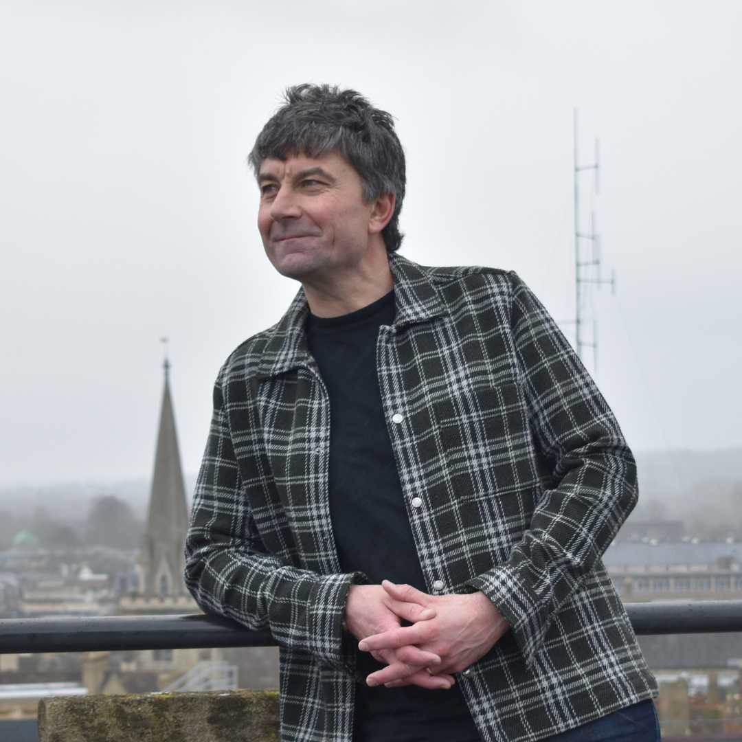 Maurice East stands in front of a view of Oxford's skyline on a cloudy day, looking off to the left. He has light-toned skin and dark, salt-and-pepper hair. He is wearing a dark plaid shirt over a tshirt.