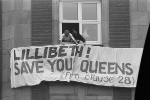 Black and white photo of two men holding a sign out of a window which reads Lilibeth! Save your Queens