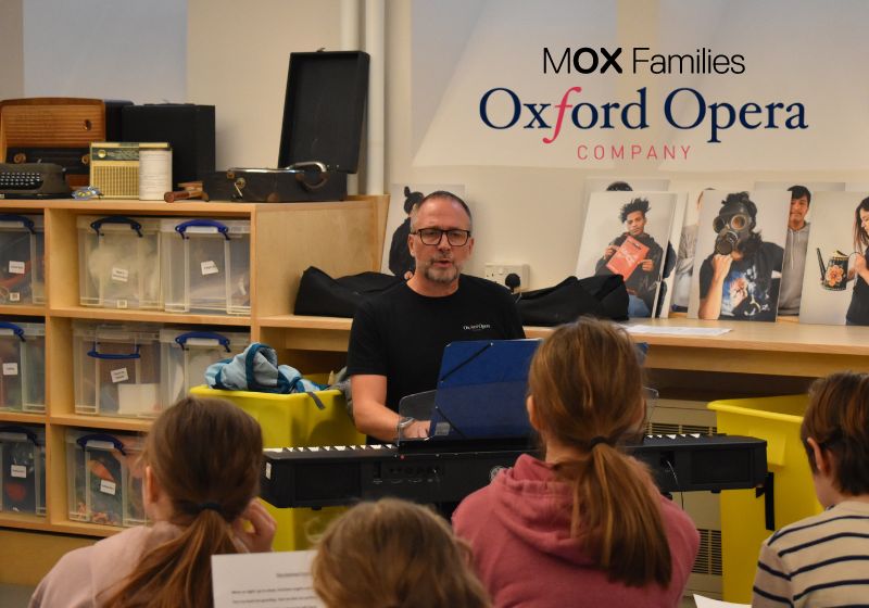 A man sits behind a keyboard and leads a singing session for several young people.