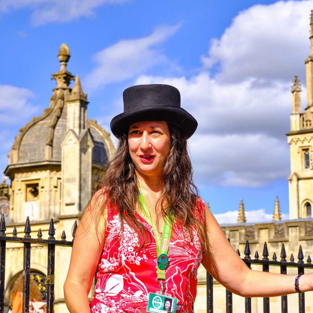 Tour guide Jess Worth stands in front of college buildings on a sunny day. She has light skin, long brown hair and is wearing a top hat and pink dress.