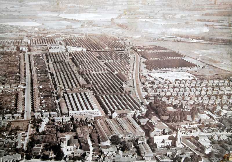 A black-and-white aerial photograph of a factory complex surrounded on one side by fields and the other by houses.