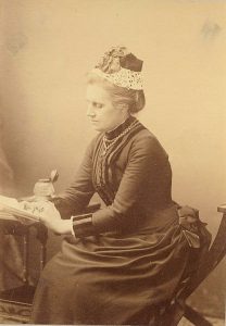 Black and white photo of a woman sitting at a desk in dark Victorian clothes.