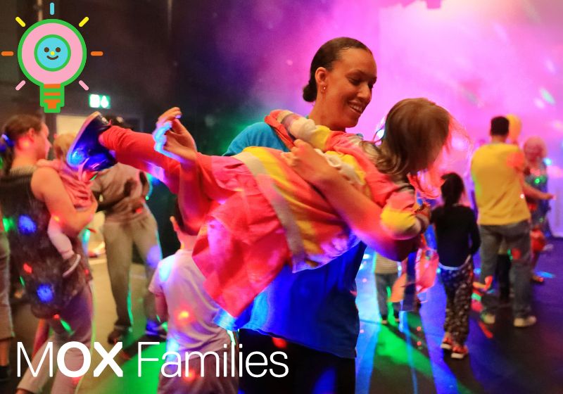 A woman with black hair cradles a child in her arms, dancing. The background of the picture shows people of all ages happily dancing in multi-coloured lights.